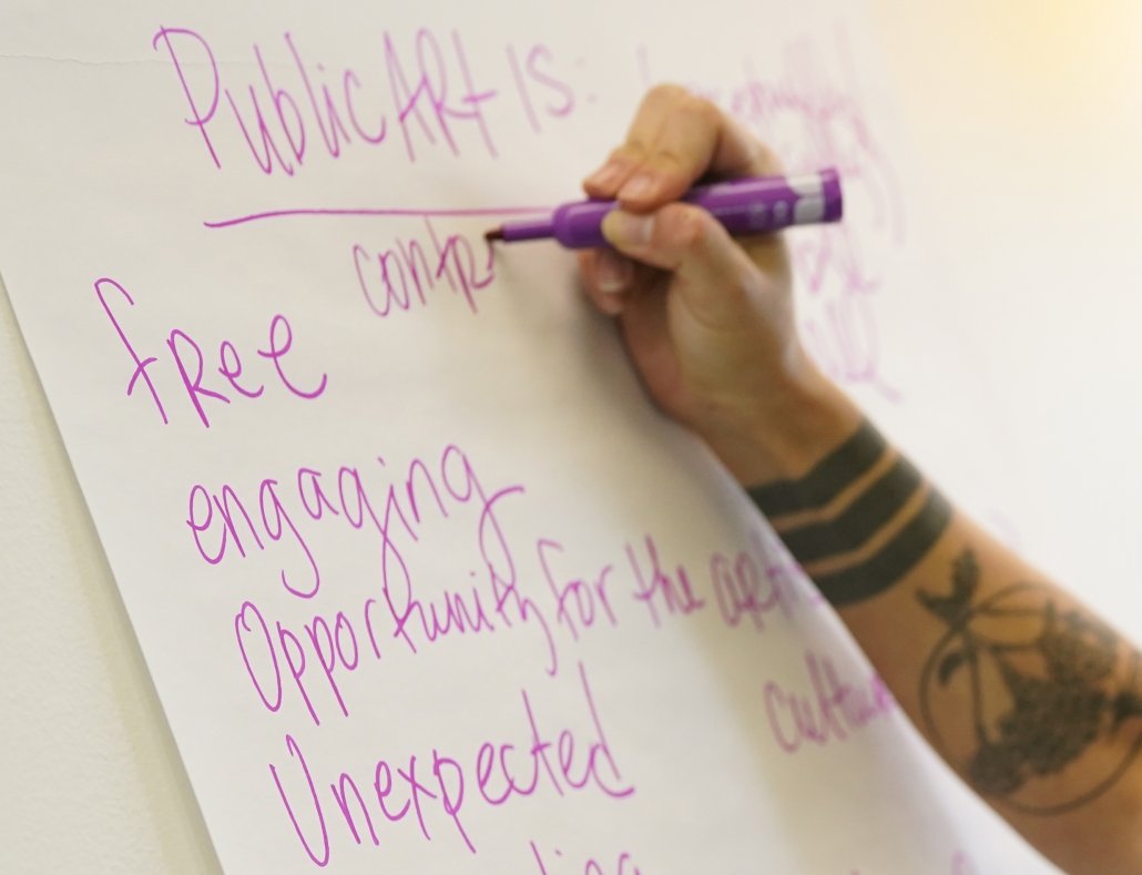a tattooed arm reaches toward a white board, the hand writing in a vibrant pink marker under a heading "Public Art Is," with words like free, engaging, opportunity, and unexpected