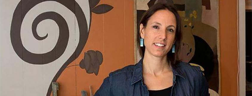 a person with shoulder length dark brown hair and dangly earrings smiles in front of a wall painted in a swirling natural toned floral motif