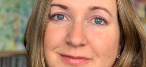 a woman with chin-length dark blonde hair and blue eyes gazes forward with a light smile