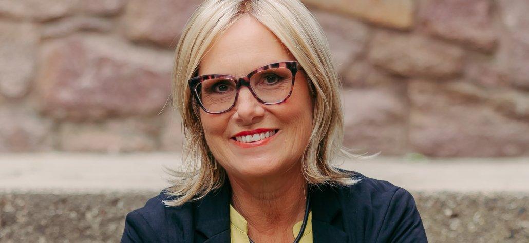 An artist wearing glasses smiles as she stands before a brick wall